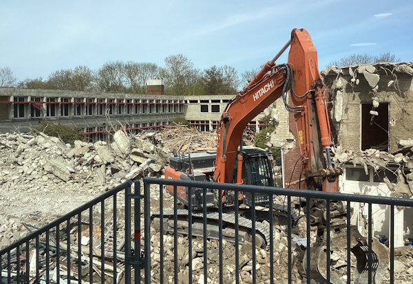 Blick von oben herab auf die Südseite des alten entkernten Schulgebäudes, dessen Südflügel komplett abgerissen wurde und so den Blick auf den mit Schotter und anderen Abbruchmaterialien gefüllten Innenhof freigibt. Im Vordergrund ein orangefarbener Bagger mit Greifarm, der die Materialien sortiert.
